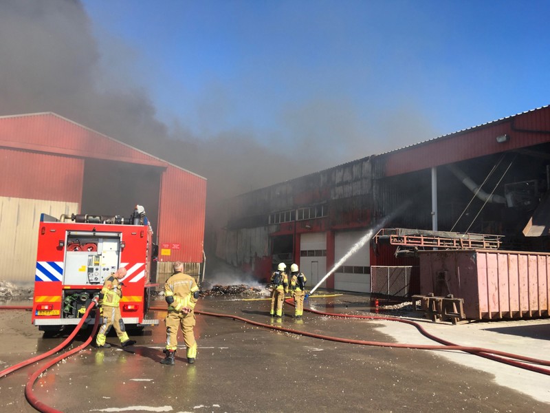 Blussende brandweermensen bij een loods waar veel rook uit komt.