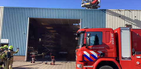 Bedrijfsloods met een brandweerauto en hoogwerker boven het dak. De brand is uit, de schade is deels zichtbaar. Nacontrole wordt uitgevoerd. 