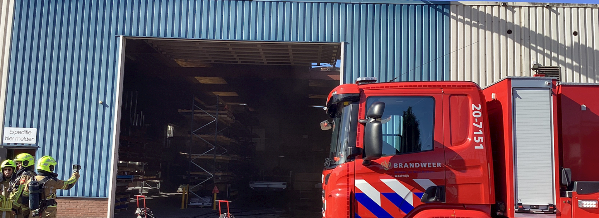 Bedrijfsloods met een brandweerauto en hoogwerker boven het dak. De brand is uit, de schade is deels zichtbaar. Nacontrole wordt uitgevoerd. 