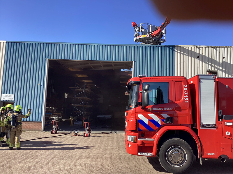 Bedrijfsloods met brandweerwagen met hoogwerker naast de loods. Geen brand meer te zien, wel de schade. 